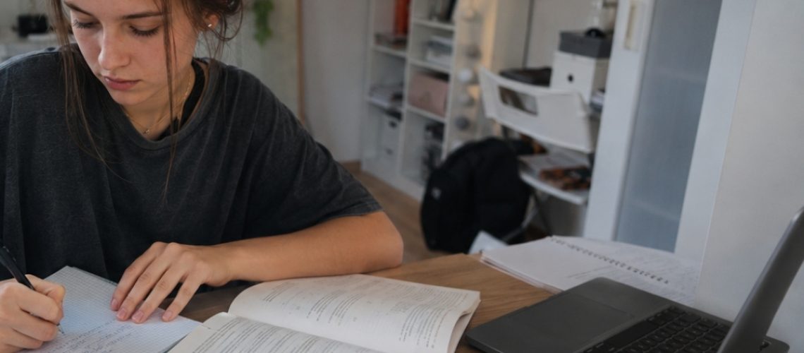 Licealistka uczy się matematyki przy biurku w pokoju, zapisując notatki obok laptopa, podręcznika, kalkulatora i arkuszy.