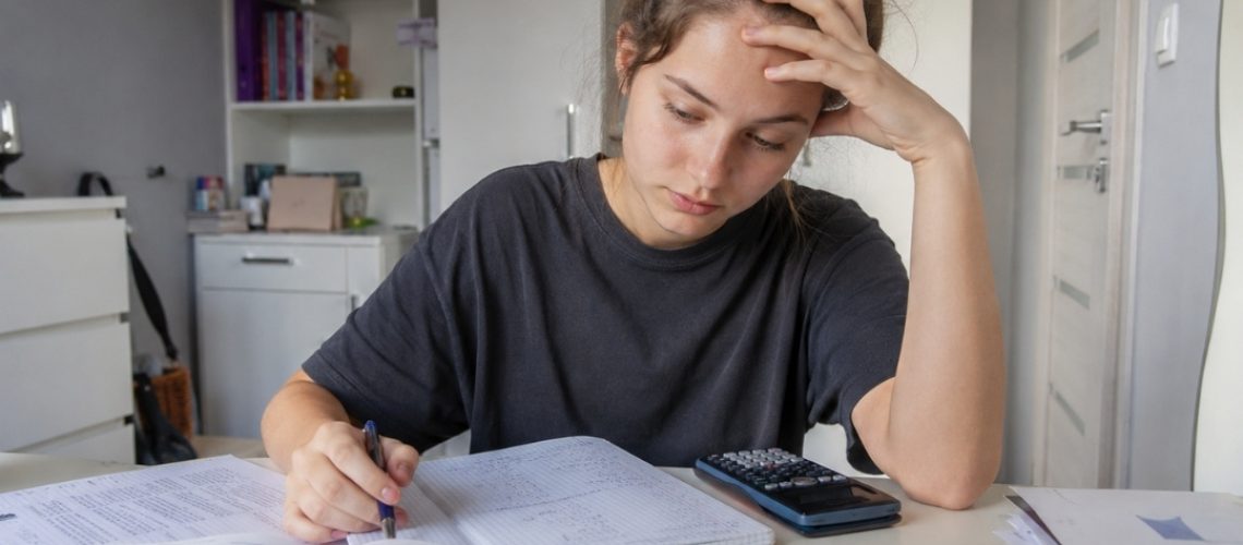 Licealistka zmaga się z zadaniem matematycznym przy biurku, zapisując obliczenia w zeszycie obok podręcznika i kalkulatora.