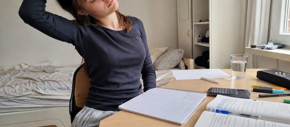 Nastolatka robi przerwę od nauki i rozciąga szyję przy biurku z zeszytem, podręcznikiem i kalkulatorem.