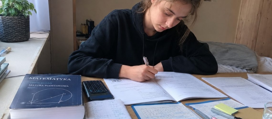 Nastolatka rozwiązuje zadania przy biurku pełnym notatek i zbioru zadań.