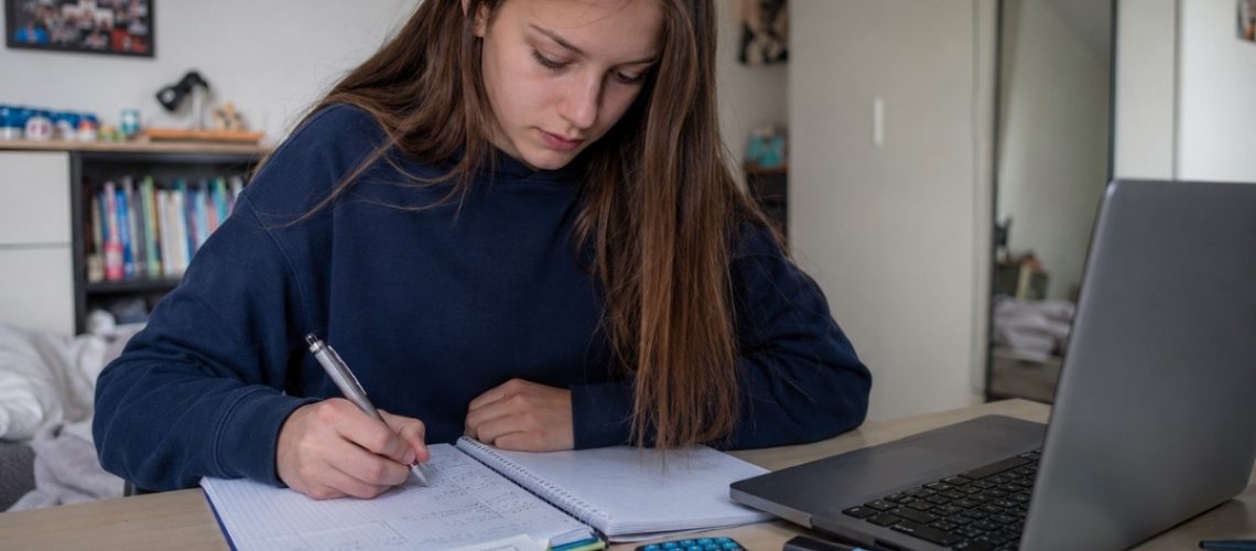 Nastolatka w wieku licealnym uczy się przy biurku, zapisując obliczenia w zeszycie obok laptopa, podręcznika i kalkulatora.