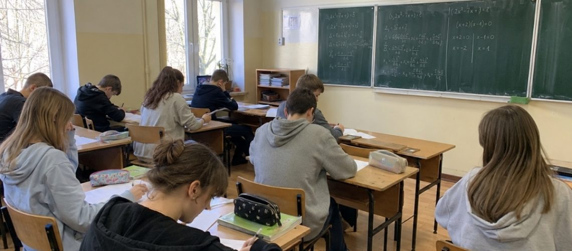 Uczniowie siedzą w ławkach w klasie i piszą w zeszytach, a na zielonej tablicy widać zapisane równania matematyczne.